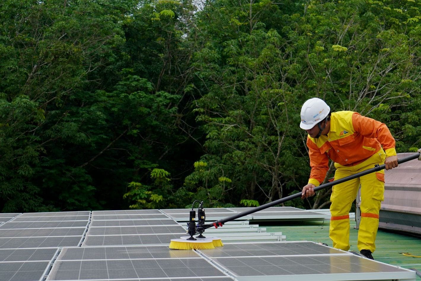 Fachkraft mit Helm reinigt auf dem Dach Solarmodule mit einer wasserführenden Teleskopbürste